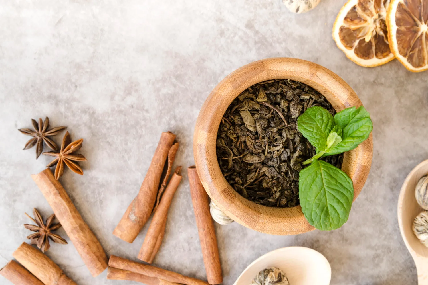 Tazón de madera con hojas de té verde y una ramita de menta fresca, rodeado de especias como canela y anís estrellado, sobre una superficie de piedra clara.