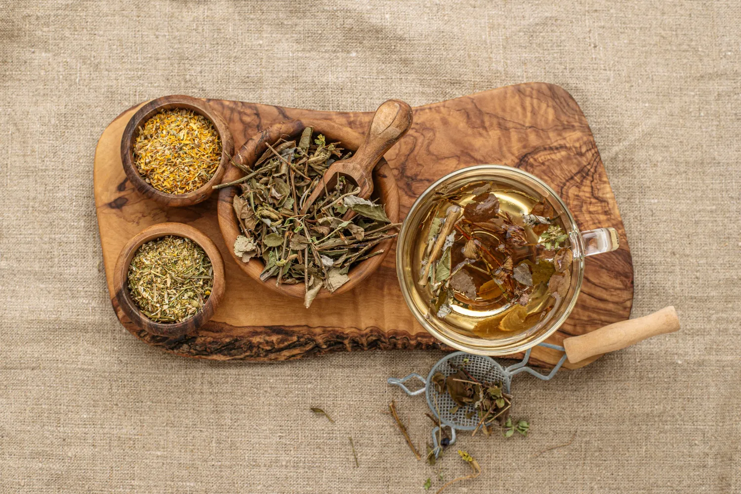 Taza de té de hierbas con infusión, rodeada de tres cuencos de madera que contienen diferentes hierbas secas, sobre una tabla de madera. Fondo de tela de lino.
