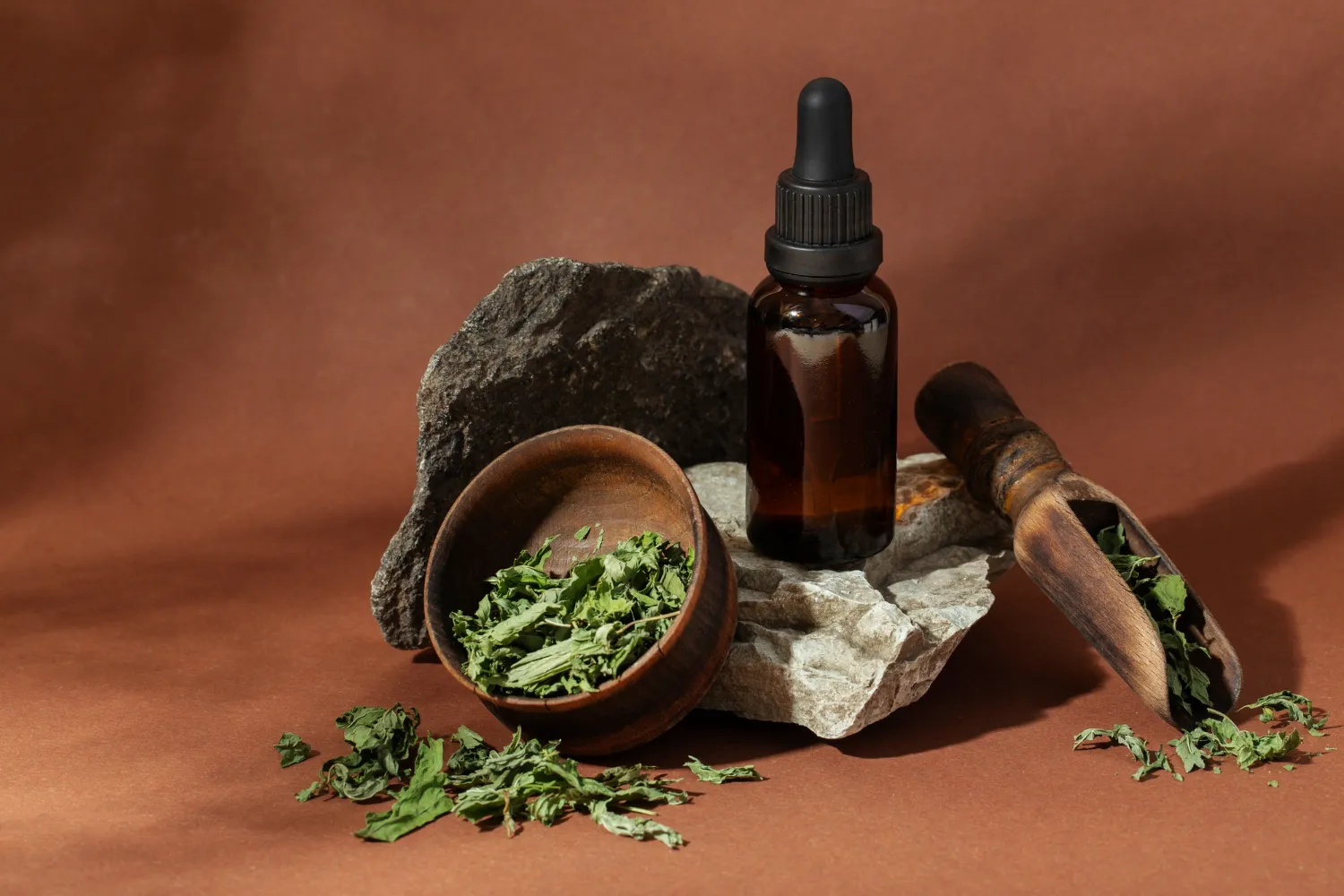 Botella de aceite esencial sobre una piedra, acompañada de un cuenco de madera con hierbas secas y una cuchara de madera, todo sobre un fondo marrón.