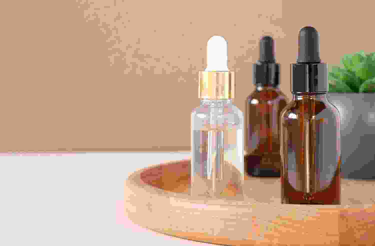 Botellas de suero facial en una bandeja de madera, con un fondo neutro. Los frascos incluyen uno transparente con tapa dorada y dos de vidrio ámbar con tapas negras. Ideal para productos de cuidado de la piel.