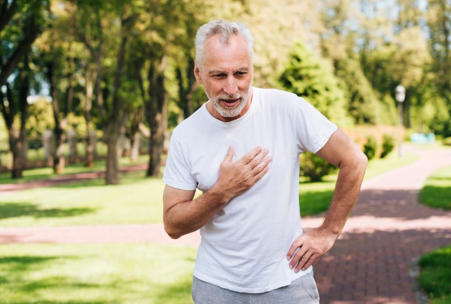 Hombre mayor con camiseta blanca sosteniéndose el pecho en un parque, mostrando signos de malestar o dolor, rodeado de naturaleza y senderos.