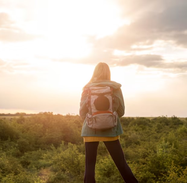 Mujer de espaldas con mochila, mirando un paisaje natural al atardecer, rodeada de vegetación.